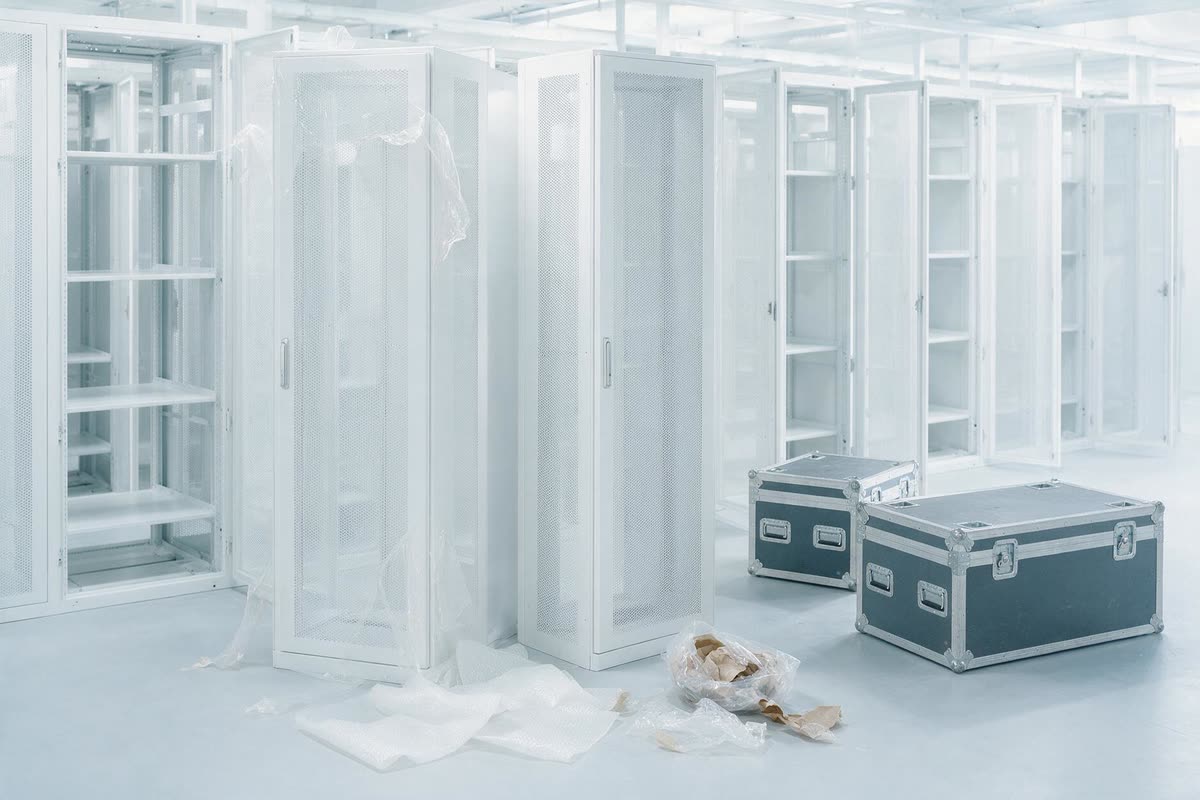 New server cabinets being positioned in a bright white data hall during a build, protective film partly removed and flight cases set aside.