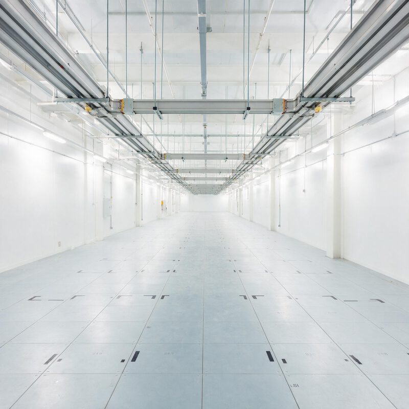 An empty white data hall before fit-out — raised floor tiles marked out for rack positions, overhead busway and containment running the length of the room.