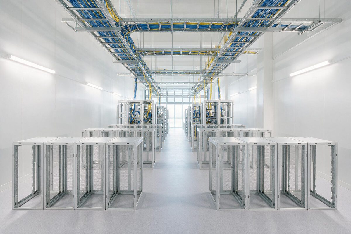 A bright data centre hall mid-build: empty aluminium rack frames in the foreground, populated racks and overhead cable trays behind